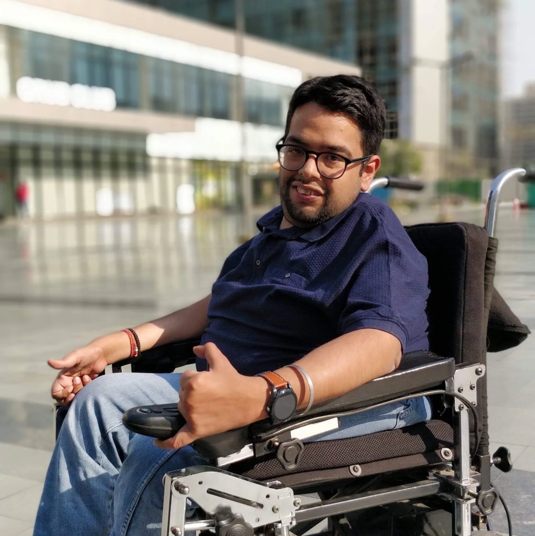Rahul in his power wheelchair on a sunny corporate campus, glass office buildings softly blurred behind him