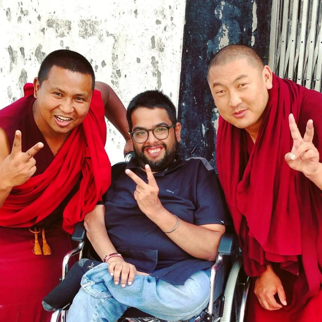 Rahul in his wheelchair, smiling and flashing a peace sign, with two monks in red robes doing the same
