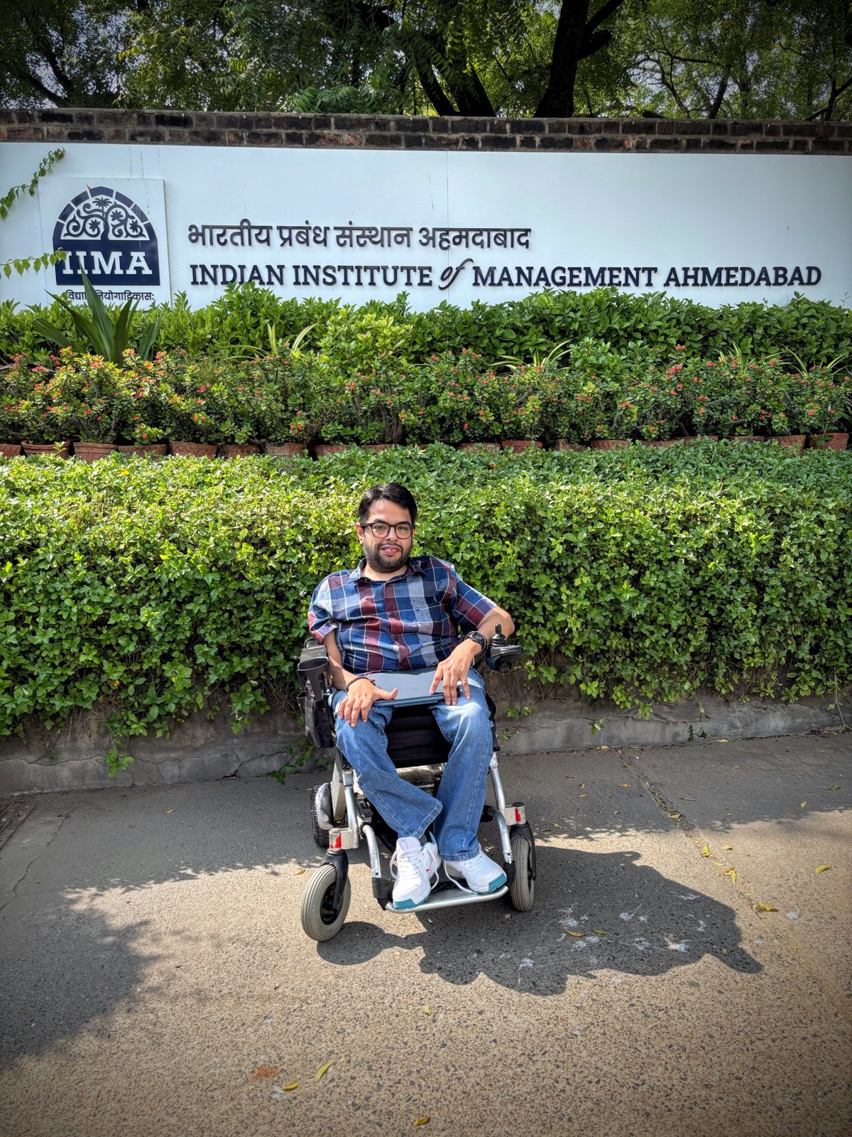 Rahul in his power wheelchair outside the main sign of the Indian Institute of Management Ahmedabad, framed by green hedges