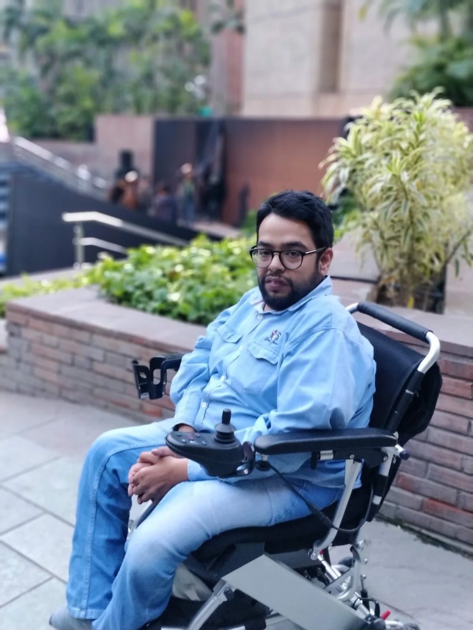 Rahul in his power wheelchair outdoors beside a planter, wearing a light blue shirt