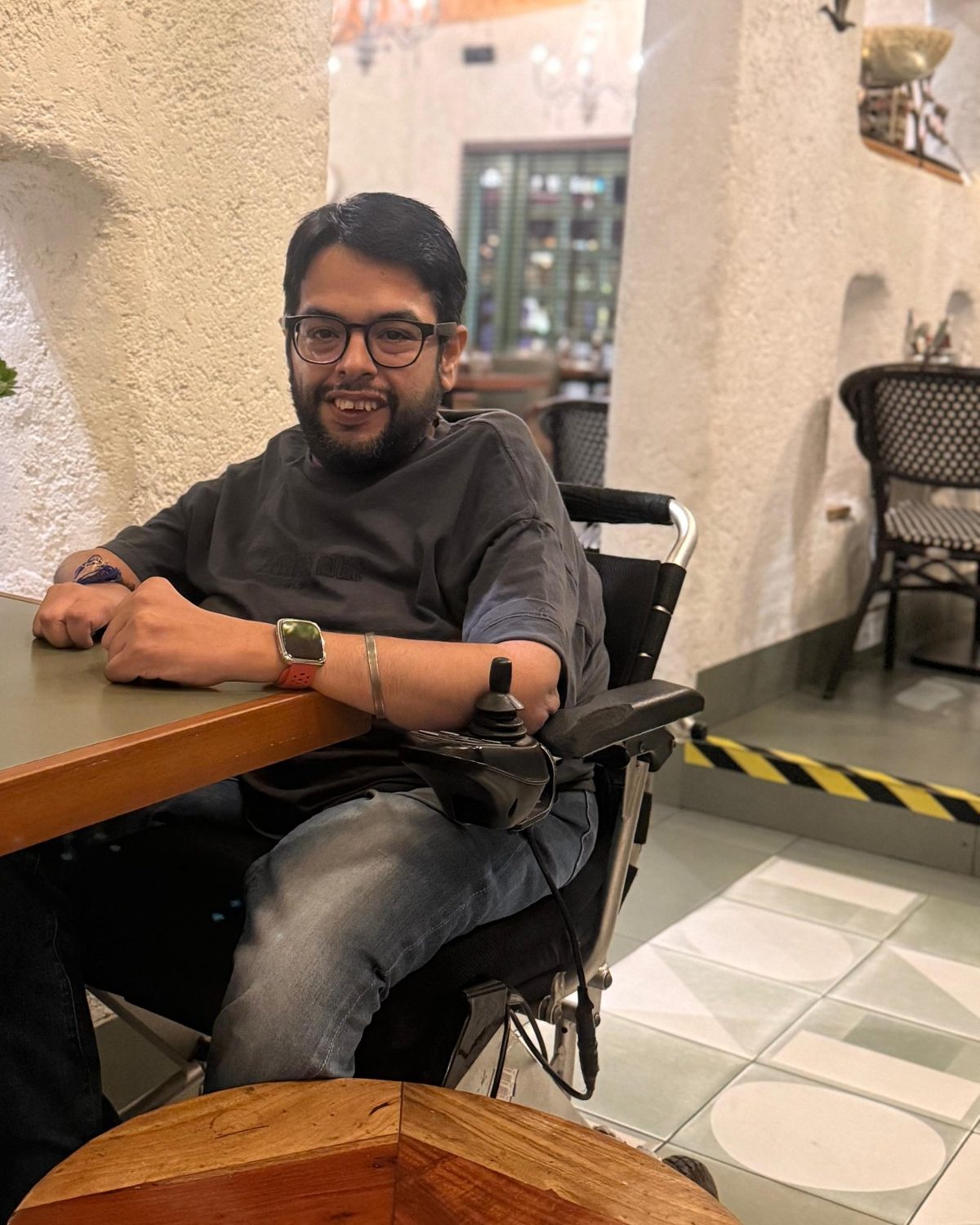 Rahul smiling at the camera from his power wheelchair, seated at a café table