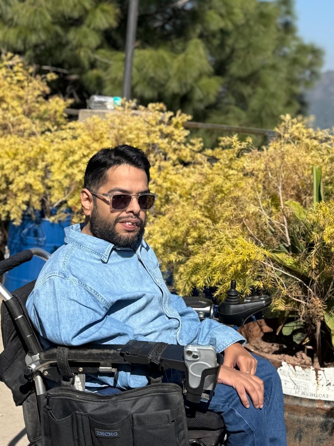 Rahul in his power wheelchair on a hillside terrace, in a denim shirt and sunglasses, with pine trees and mountains behind him