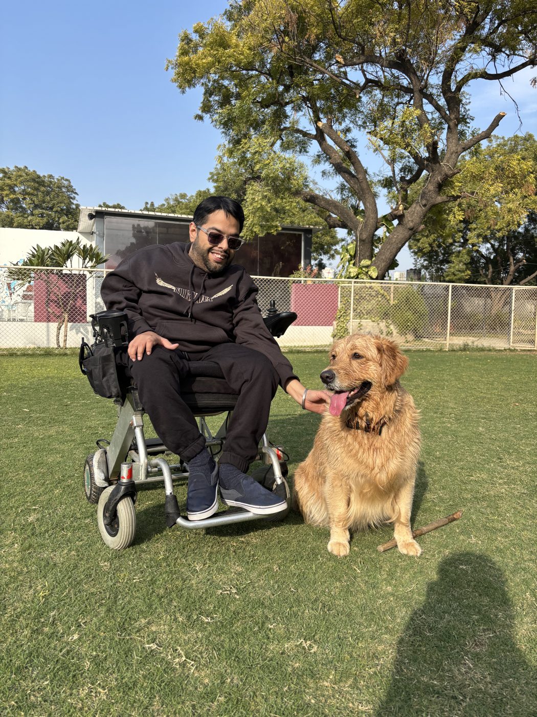 Rahul smiling in his power wheelchair on a sunny lawn, one hand resting on Zoe, his golden retriever, sitting beside him